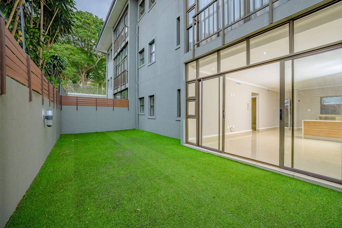 A private rear garden area featuring artificial turf, a secure boundary wall, and large sliding glass doors providing access to the apartment interior.