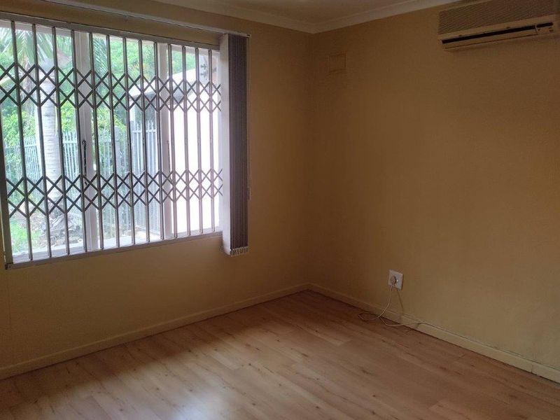 An empty bedroom featuring light laminate flooring, a wall-mounted air conditioning unit, and a window fitted with a security gate and vertical blinds.