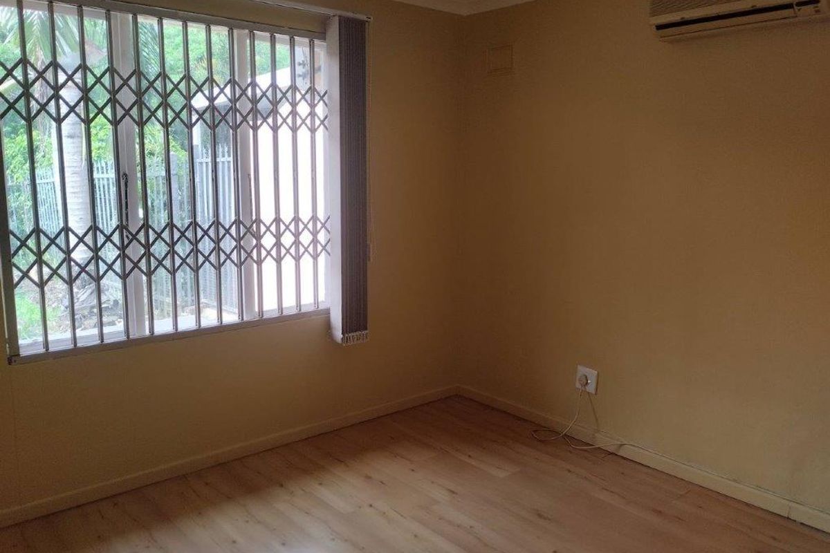 An empty bedroom featuring light laminate flooring, a wall-mounted air conditioning unit, and a window fitted with a security gate and vertical blinds.