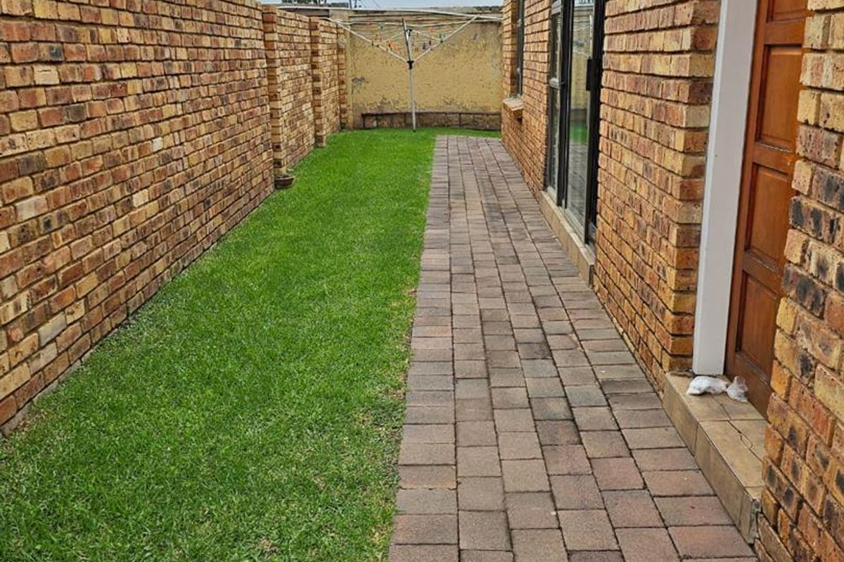 A side garden area featuring a paved walkway, well-maintained lawn, and face brick boundary walls, providing access to the home via a sliding glass door.