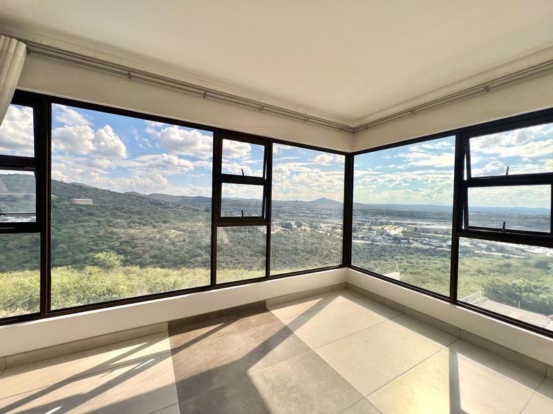A bright, empty living area featuring expansive corner windows that offer panoramic views of the surrounding landscape and hills.