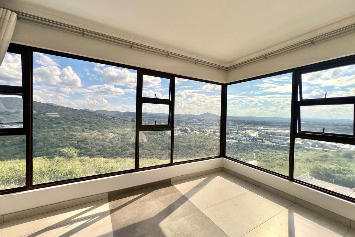 A bright, empty living area featuring expansive corner windows that offer panoramic views of the surrounding landscape and hills.