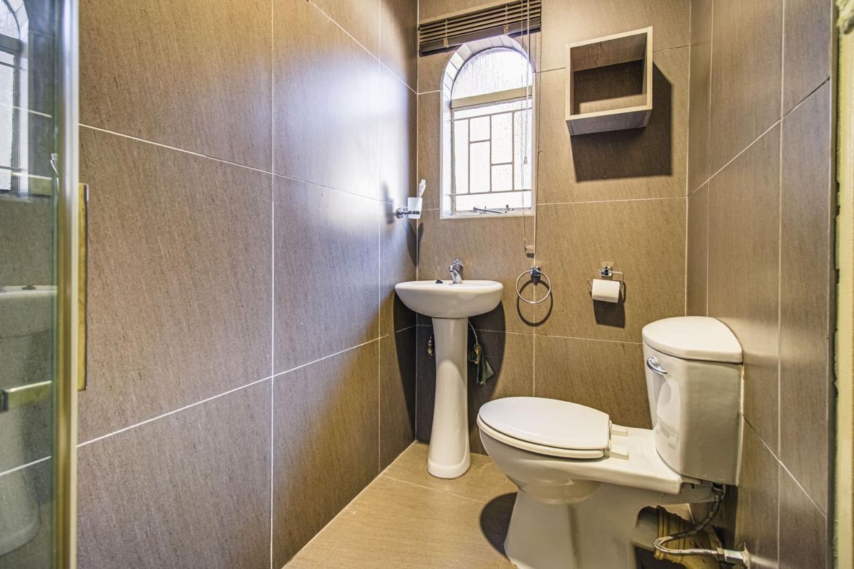 A compact bathroom featuring full-height wall tiling, a pedestal basin, a standard toilet, and an arched window with a fitted blind.
