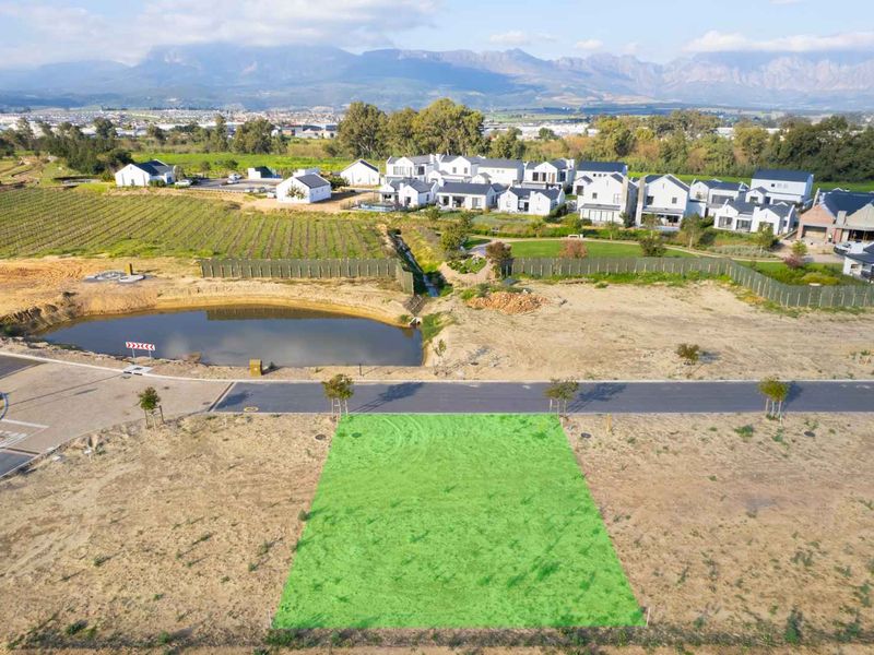 Aerial view of a vacant residential plot in a developed estate, featuring road access and proximity to a scenic pond and mountain views.