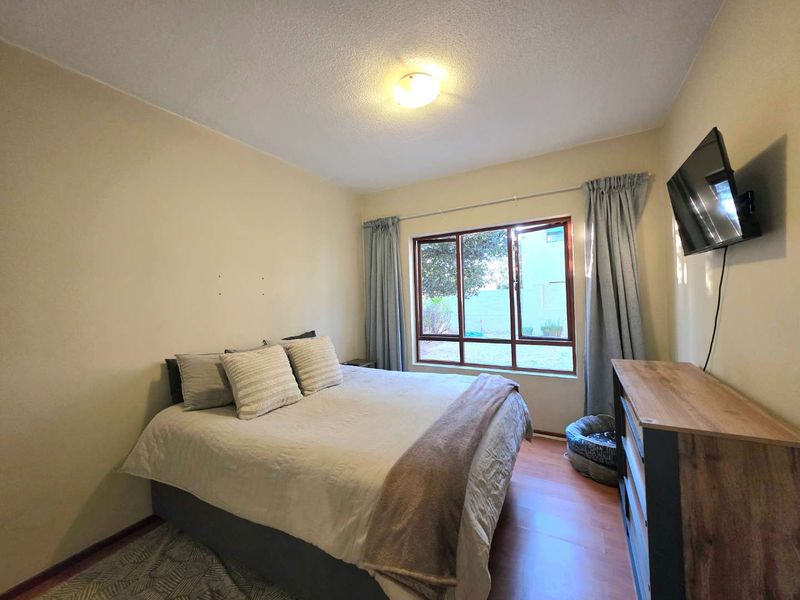 A functional bedroom featuring laminate flooring, a large window providing natural light, and a neutral wall finish.