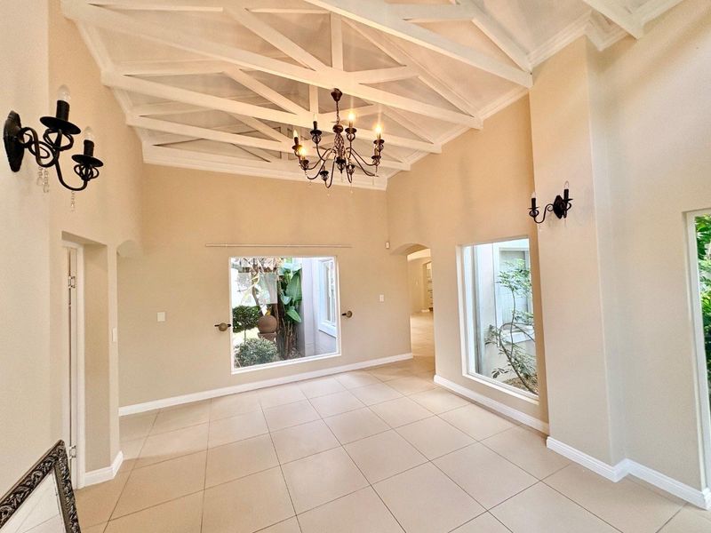 Spacious living area featuring a high vaulted ceiling with exposed beams, neutral tiled flooring, and large windows overlooking a private garden courtyard.
