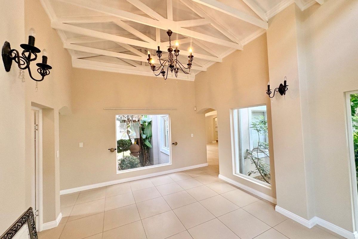 Spacious living area featuring a high vaulted ceiling with exposed beams, neutral tiled flooring, and large windows overlooking a private garden courtyard.