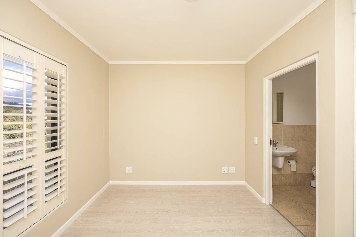 A bright, empty bedroom featuring light wood-look flooring, plantation shutters, and an en-suite bathroom with tiled finishes.
