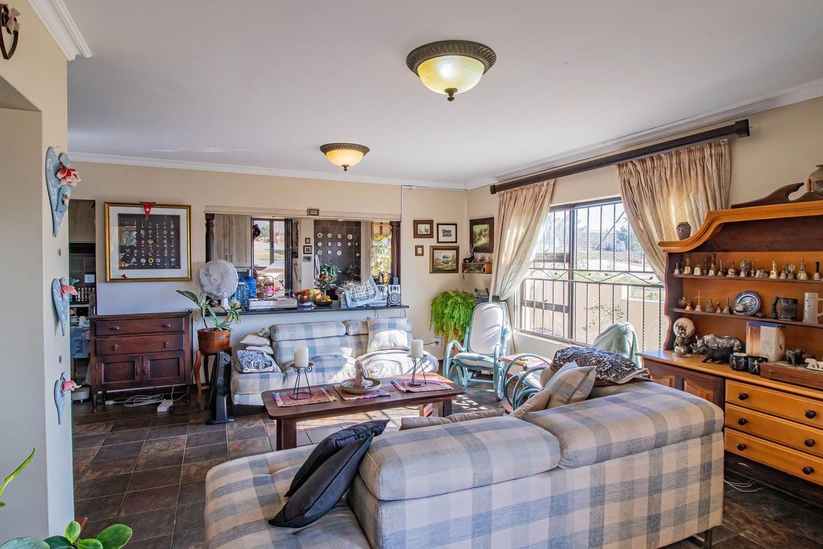 A comfortable living room featuring tiled flooring, large windows with security bars, and a pass-through window connecting to the kitchen area.