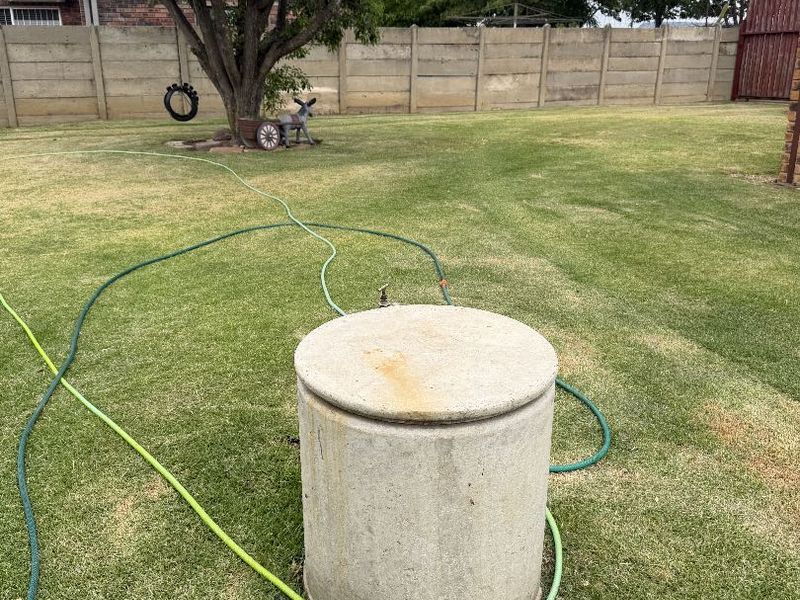 A spacious backyard featuring a lawn, mature tree, concrete boundary wall, and a central concrete septic tank cover with an integrated water tap.
