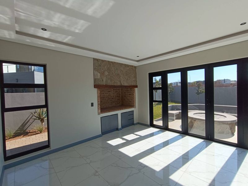 Bright living area featuring large aluminium stack doors opening to the patio, built-in braai with stone cladding, and modern marble-look tiled flooring.