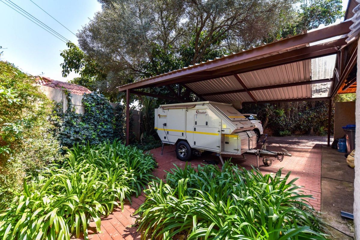 A paved rear outdoor area featuring a covered carport structure, established garden beds, and boundary walling.