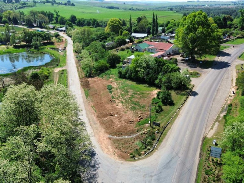 Aerial view of a vacant commercial land parcel featuring road frontage, mature trees, and proximity to a scenic water body in a rural setting.