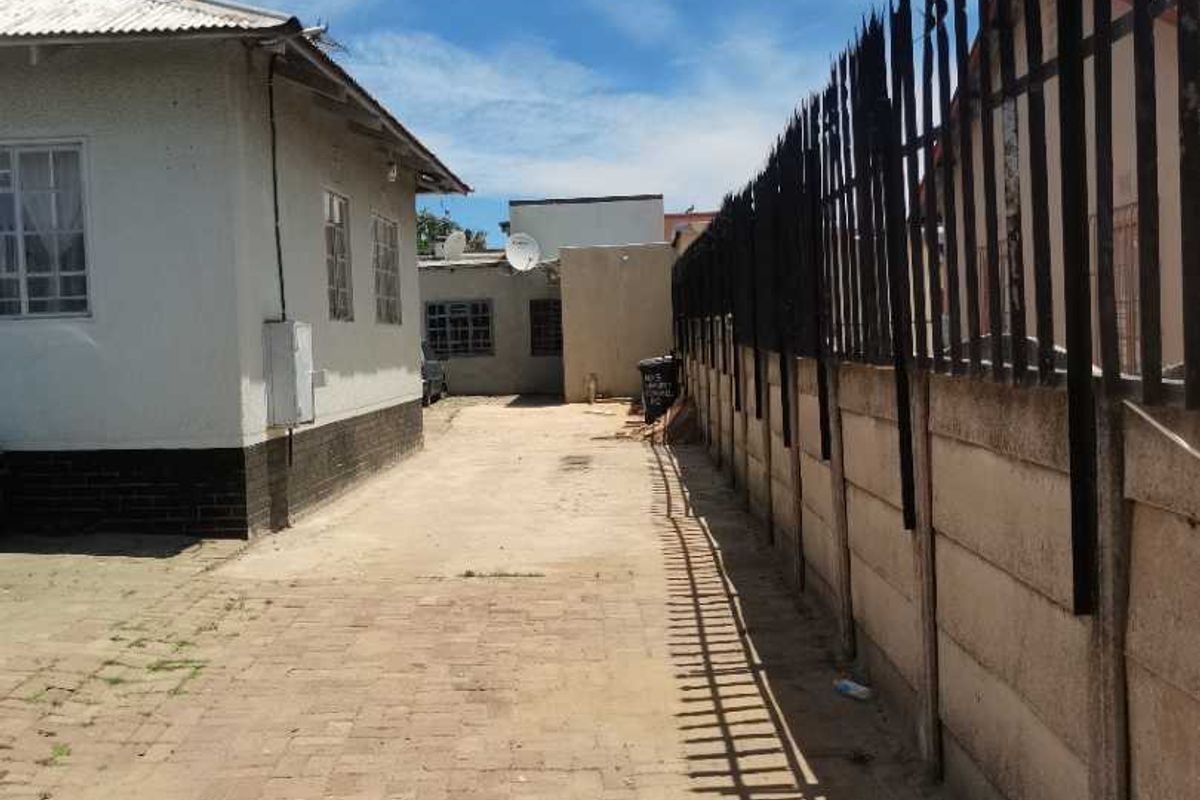 A side exterior view showing a paved driveway, a precast concrete boundary wall with security spikes, and the side elevation of the house.