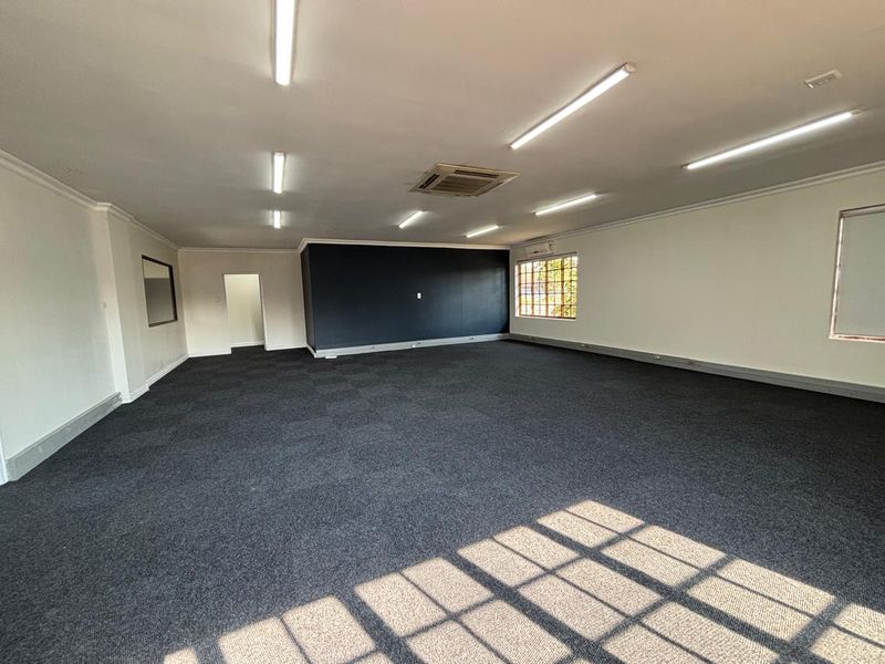 Spacious, open-plan office area featuring dark carpet tiles, recessed air conditioning, and ample natural light through grid windows.