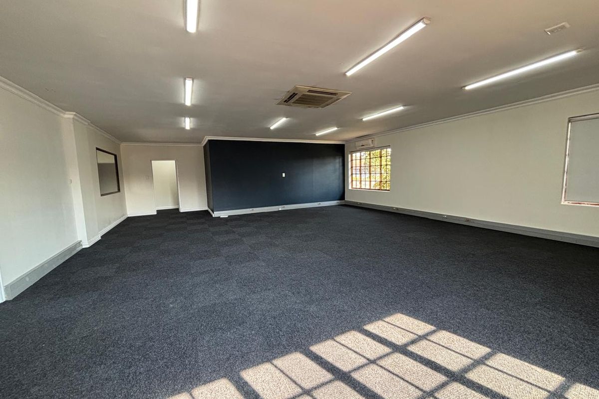 Spacious, open-plan office area featuring dark carpet tiles, recessed air conditioning, and ample natural light through grid windows.