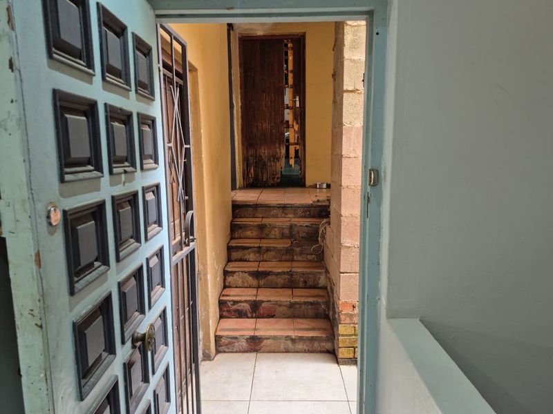A view through a panelled door into a hallway featuring a tiled staircase and security gate, showing signs of wear and maintenance needs.