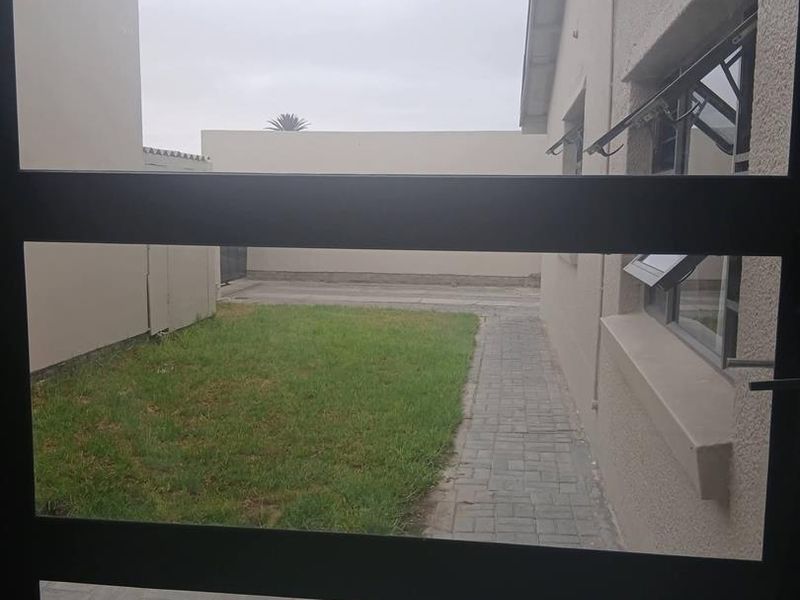 View of the rear garden area featuring a paved walkway, lawn, and boundary wall, captured through a window frame.