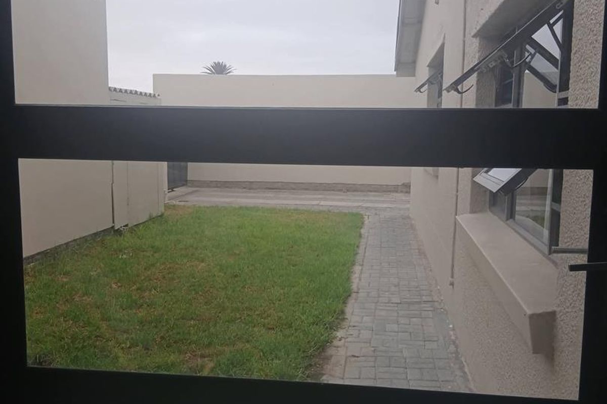 View of the rear garden area featuring a paved walkway, lawn, and boundary wall, captured through a window frame.