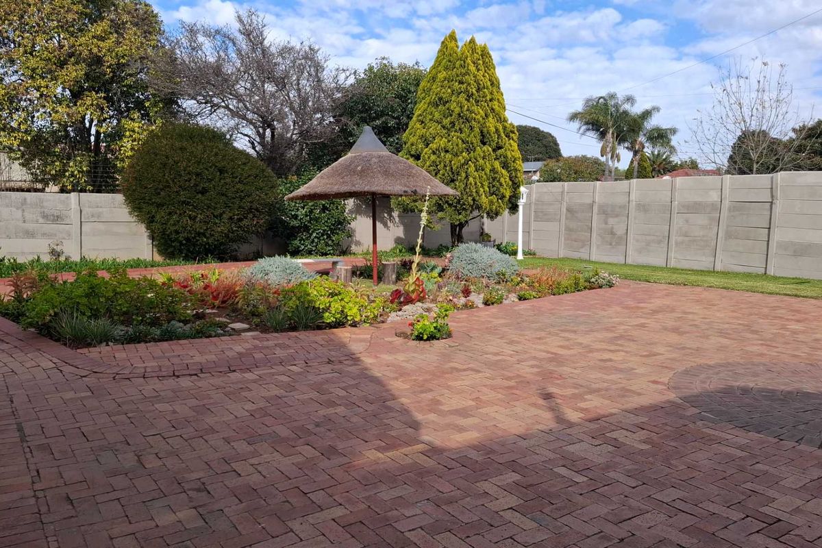 A well-maintained rear garden featuring extensive brick paving, a central landscaped garden bed, and a thatched lapa structure enclosed by concrete perimeter walling.