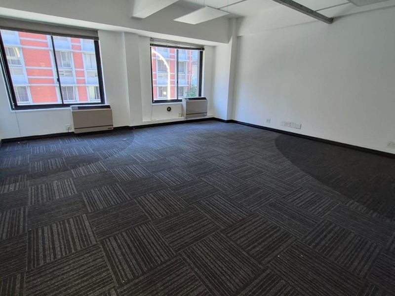 A spacious, empty office suite featuring dark carpet tiles, suspended ceiling panels, and integrated HVAC units with large windows providing natural light.