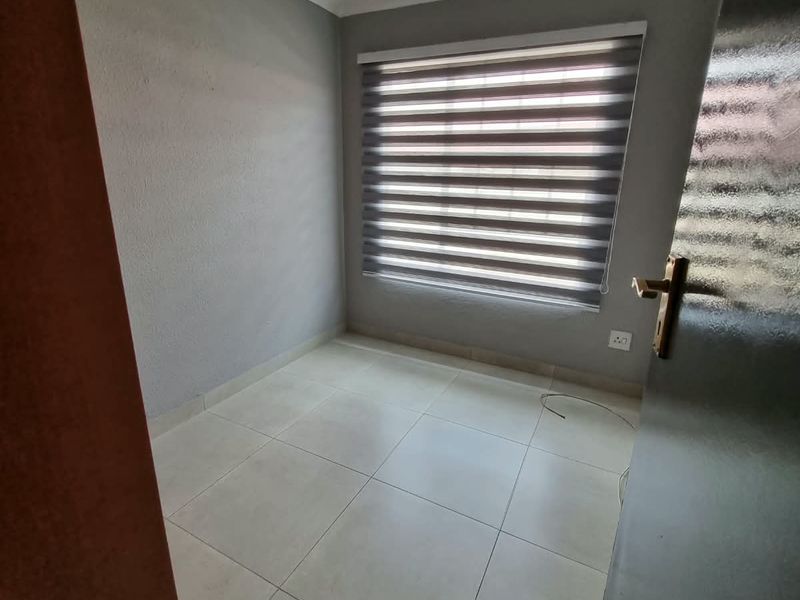 A clean, empty bedroom featuring neutral grey walls, tiled flooring, recessed downlighting, and modern roller blinds.