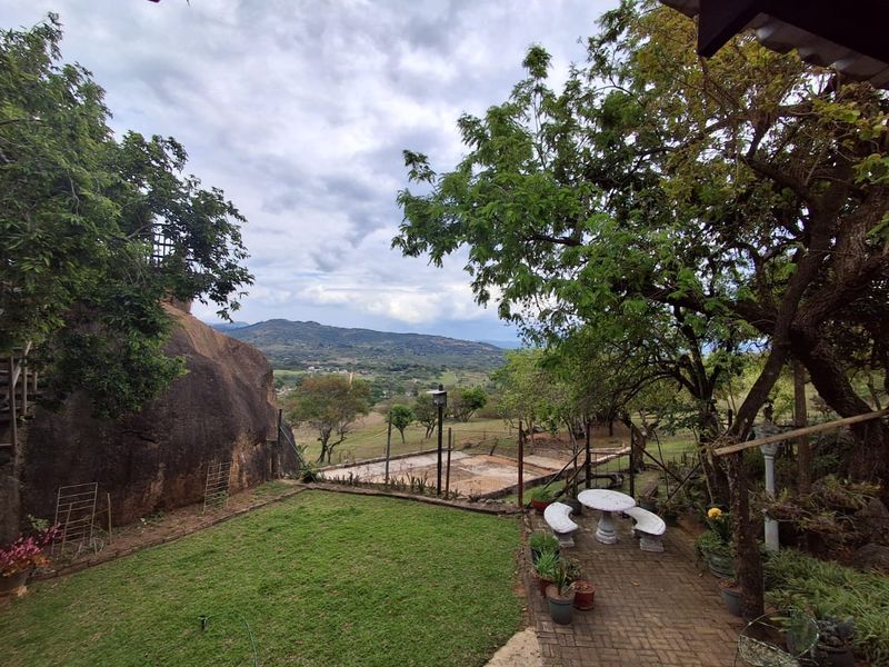 Scenic outdoor view overlooking a rural landscape, featuring a paved patio area, established trees, and a natural rock formation.