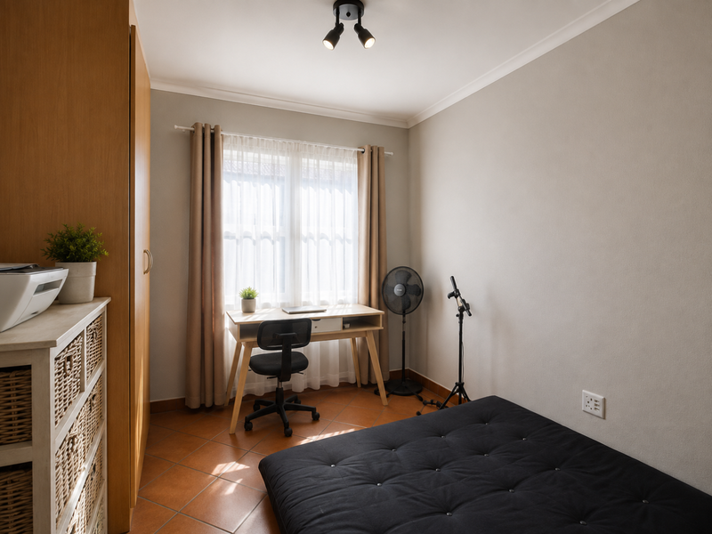 A functional bedroom featuring terracotta floor tiles, a built-in wardrobe, and a window with natural light, providing a versatile space for rest or study.