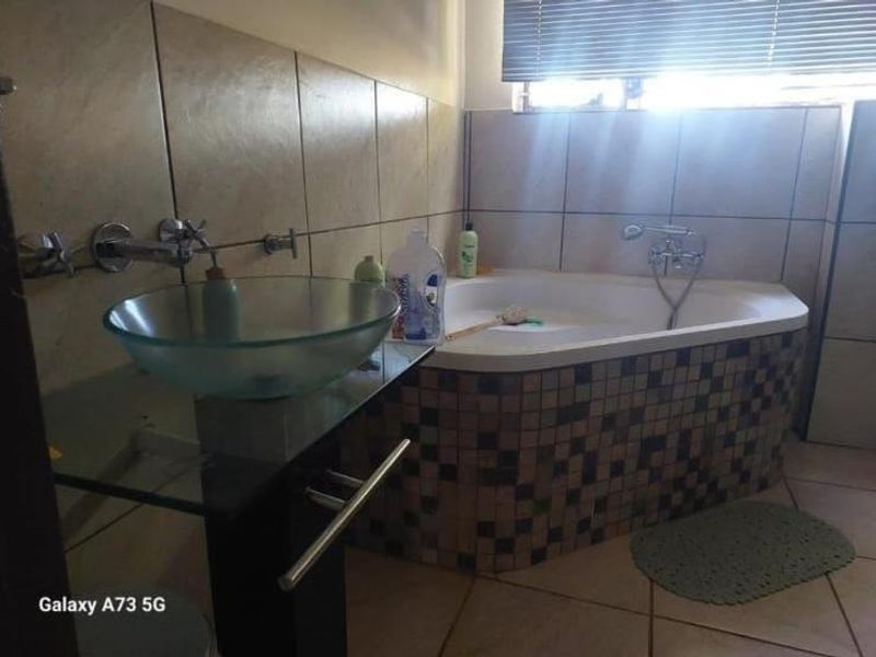 A bathroom featuring a corner bathtub with mosaic tiling, a glass vessel sink, and wall-mounted fixtures, finished with neutral wall and floor tiles.