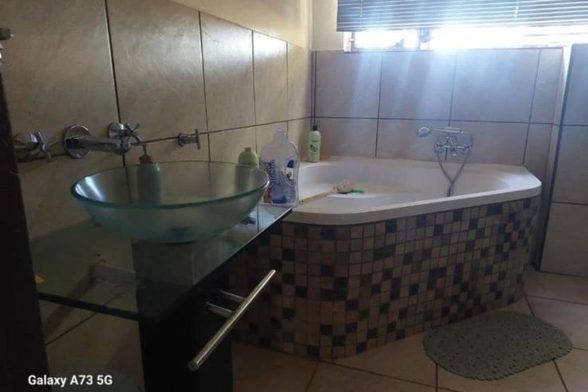 A bathroom featuring a corner bathtub with mosaic tiling, a glass vessel sink, and wall-mounted fixtures, finished with neutral wall and floor tiles.