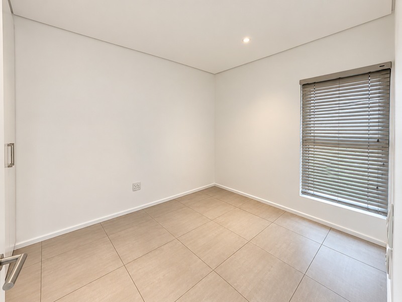 A bright, empty bedroom featuring neutral tiled flooring, recessed downlighting, and fitted venetian blinds, offering a clean and versatile space.