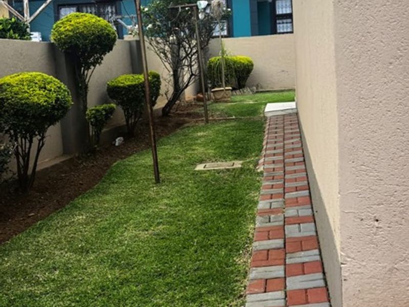 A side garden area featuring a paved walkway along the house exterior, a well-maintained lawn, and boundary walling.