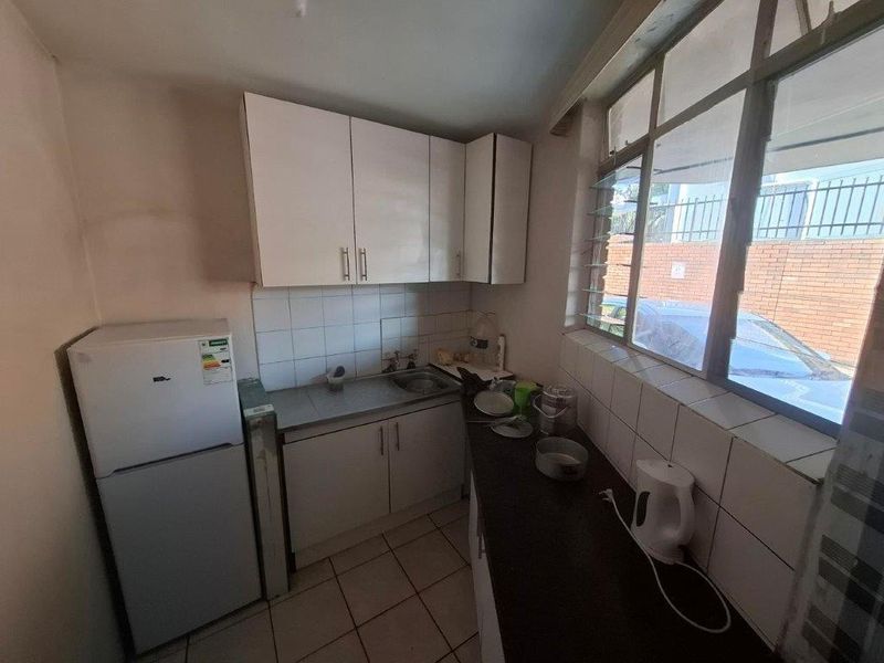 Compact kitchen featuring white cabinetry, stainless steel sink, tiled splashback, and tiled flooring, with natural light provided by large louvre windows.