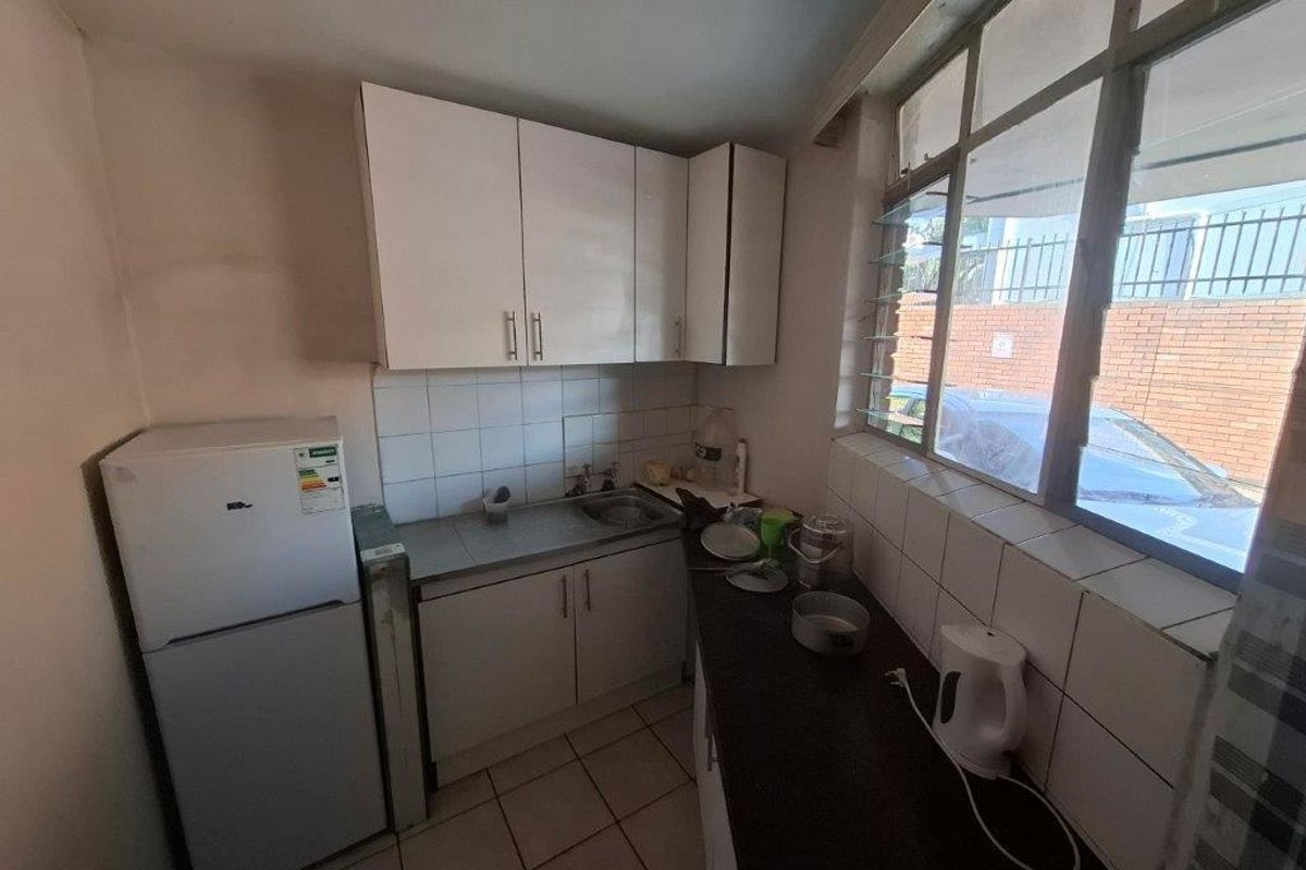 Compact kitchen featuring white cabinetry, stainless steel sink, tiled splashback, and tiled flooring, with natural light provided by large louvre windows.