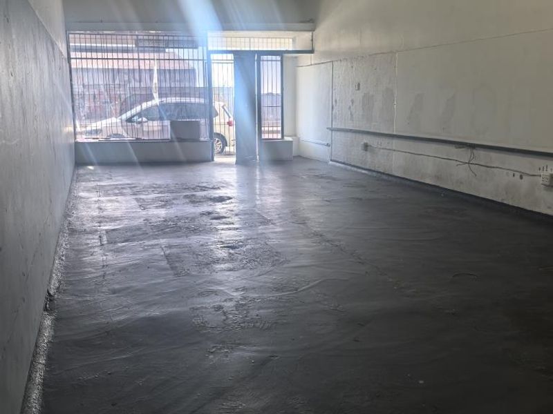 An empty, open-plan commercial space featuring concrete flooring, security bars on windows, and basic fluorescent lighting, suitable for retail or office use.