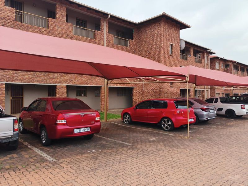 Exterior view of a multi-story face brick apartment complex featuring paved parking bays with shade cloth carports.