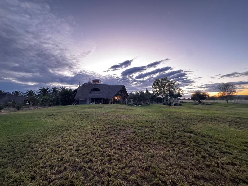 A wide-angle view of a farm property featuring a distinctive thatched-roof building set against a sunset sky and expansive lawn.