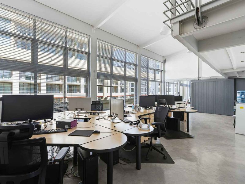A bright, modern open-plan office space featuring polished concrete floors, large industrial windows with venetian blinds, and an exposed steel mezzanine structure.