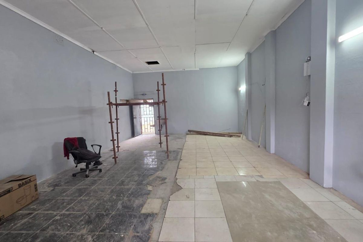 An empty open-plan commercial space featuring mixed tiled flooring, a suspended ceiling, and a security gate, currently undergoing maintenance or renovation.