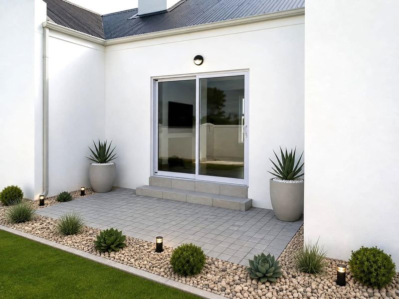 A clean, modern paved patio area featuring a sliding glass door entrance, exterior wall lighting, and neatly landscaped pebble garden beds.