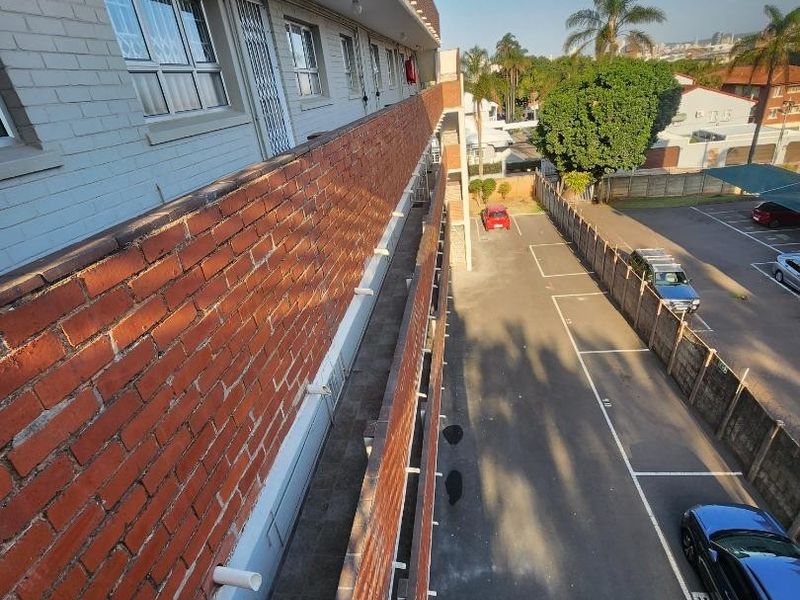 Exterior view of the apartment building showing the rear facade, access walkway, metal railings, and the adjacent paved parking area.