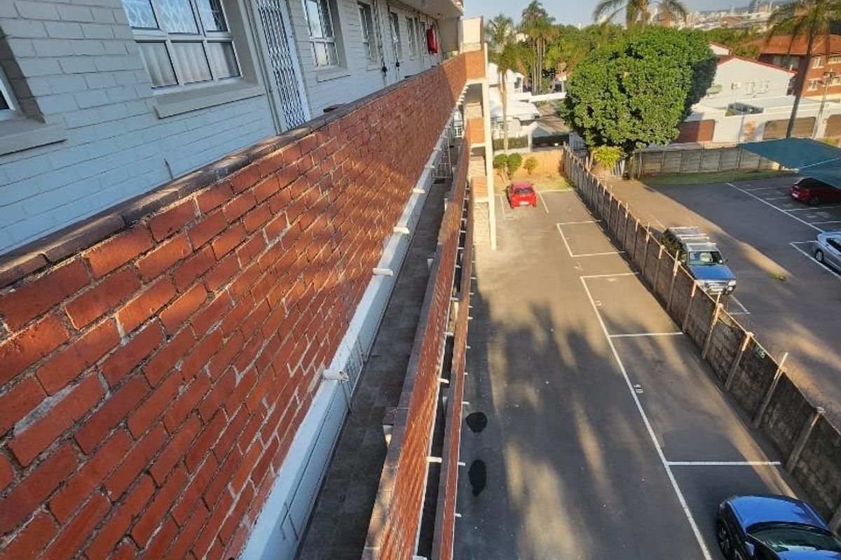Exterior view of the apartment building showing the rear facade, access walkway, metal railings, and the adjacent paved parking area.