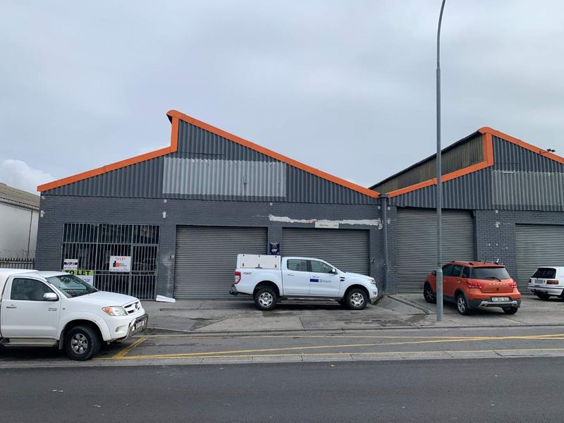 Front elevation of a commercial retail property featuring multiple roller shutter doors, brick facade, and corrugated metal roofing.