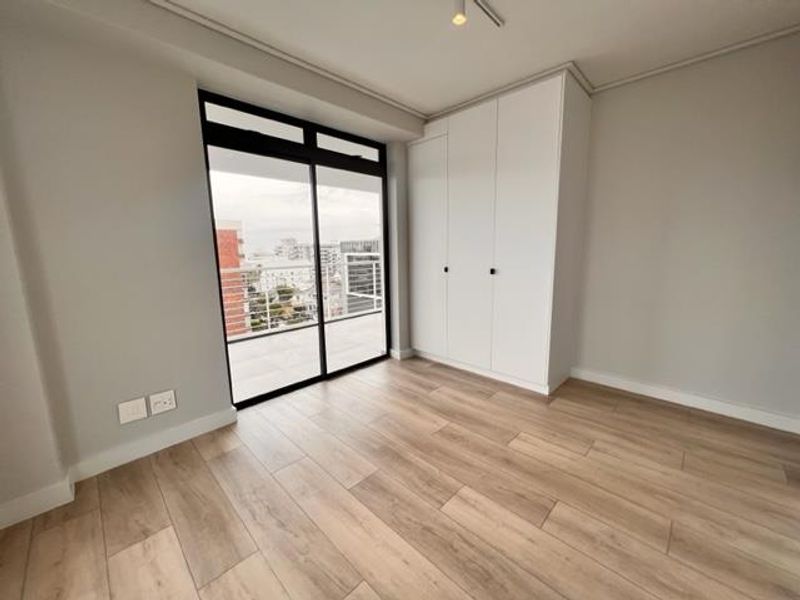 A bright, modern bedroom featuring wood-look tile flooring, a built-in wardrobe, and a sliding glass door leading to an outdoor balcony.