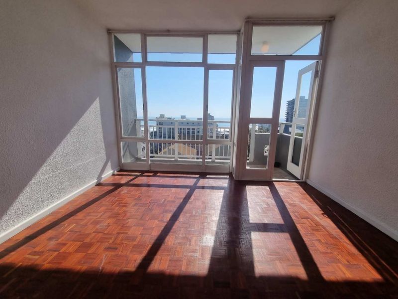 An empty living room featuring classic parquet flooring, large windows providing natural light, and direct access to an outdoor balcony area.