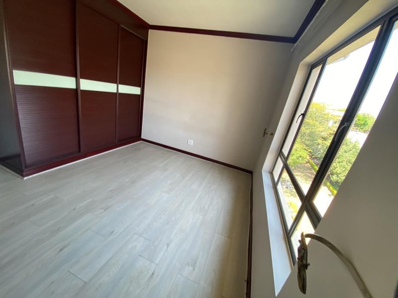 A bright, empty bedroom featuring light-toned wood-look flooring, dark wood-finish built-in sliding door wardrobes, and large windows providing natural light.