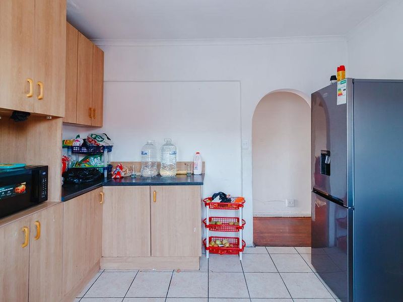 A functional kitchen featuring light wood-tone cabinetry, dark laminate countertops, and tiled flooring, leading through an arched doorway to adjacent living spaces.