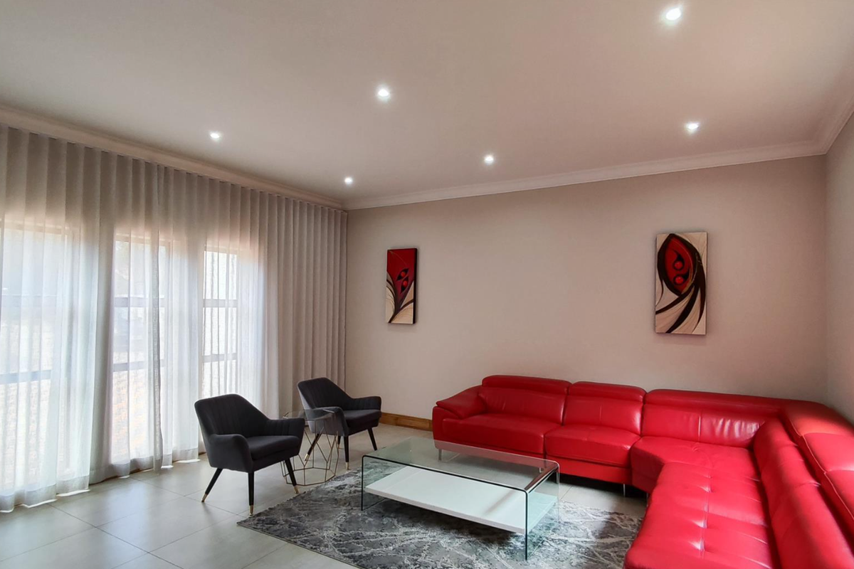 A bright and spacious living room featuring recessed ceiling lighting, large windows with floor-to-ceiling curtains, and neutral tiled flooring.