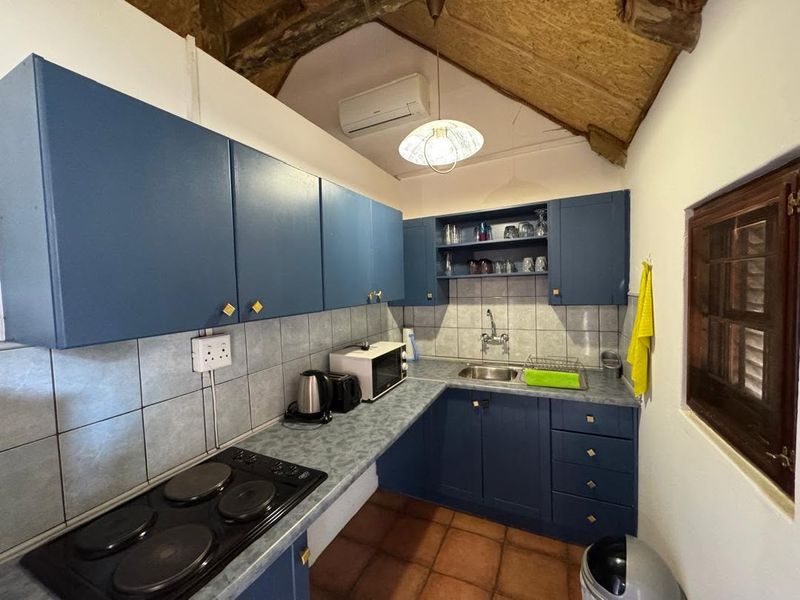 A compact kitchen featuring blue cabinetry, an electric hob, tiled backsplash, and exposed timber ceiling beams with terracotta flooring.