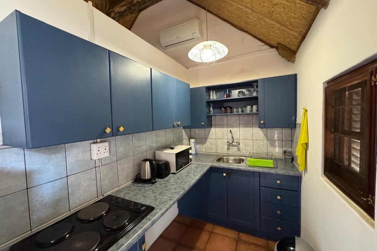 A compact kitchen featuring blue cabinetry, an electric hob, tiled backsplash, and exposed timber ceiling beams with terracotta flooring.